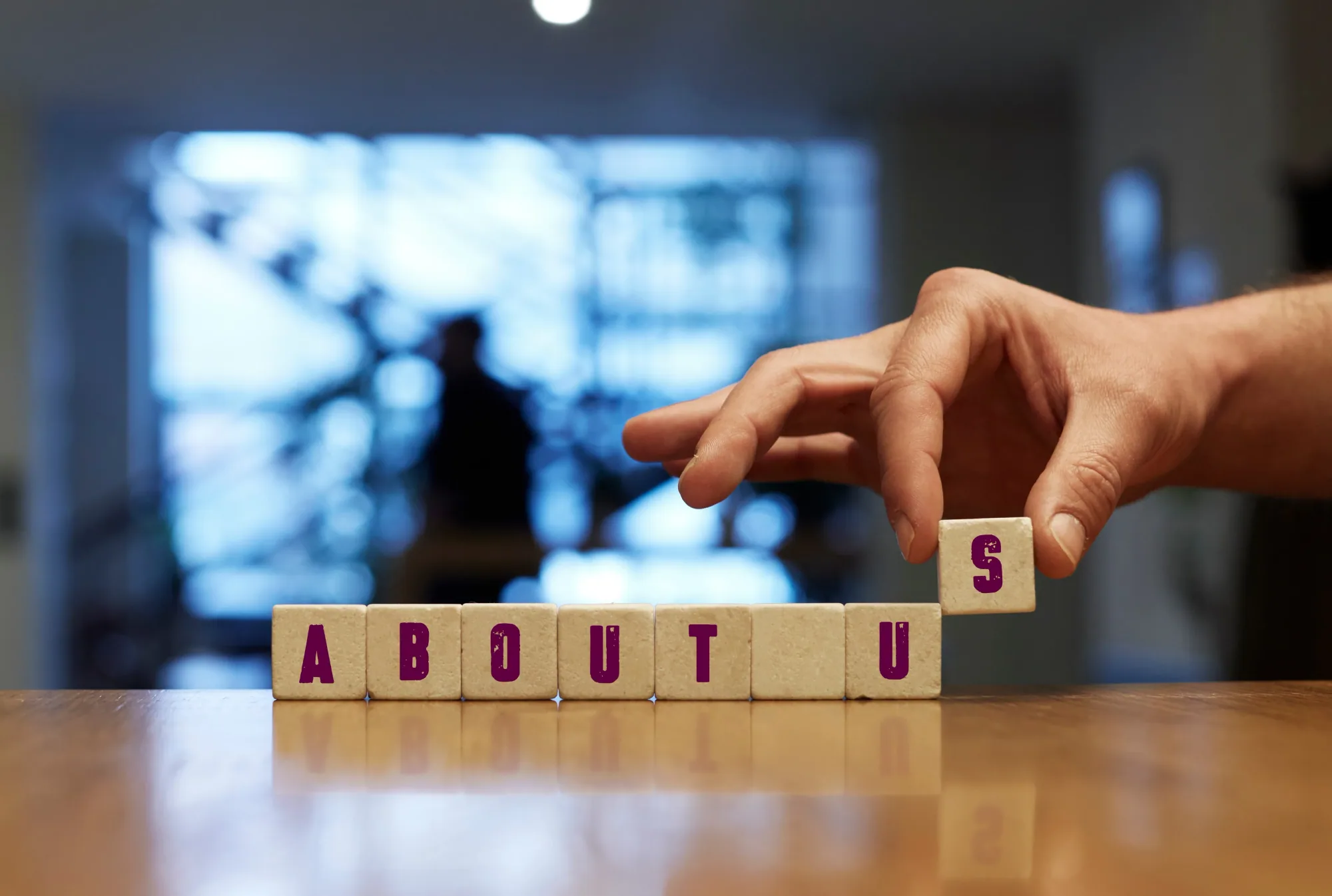 A hand placing the final lettered tile to complete the words ‘About Us’ on a wooden table.